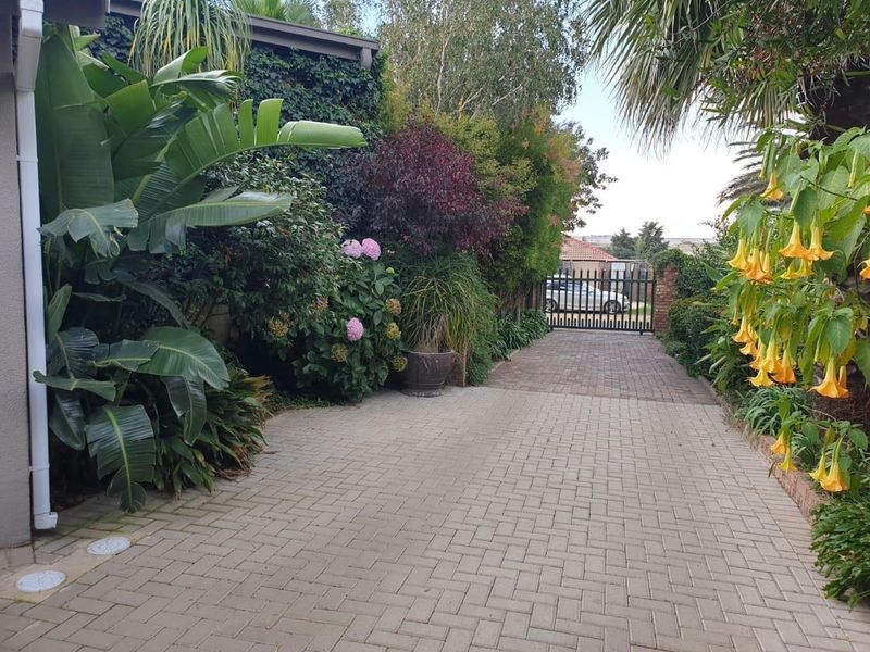 A paved driveway leading to a secure metal gate, flanked by lush established garden beds and mature greenery.