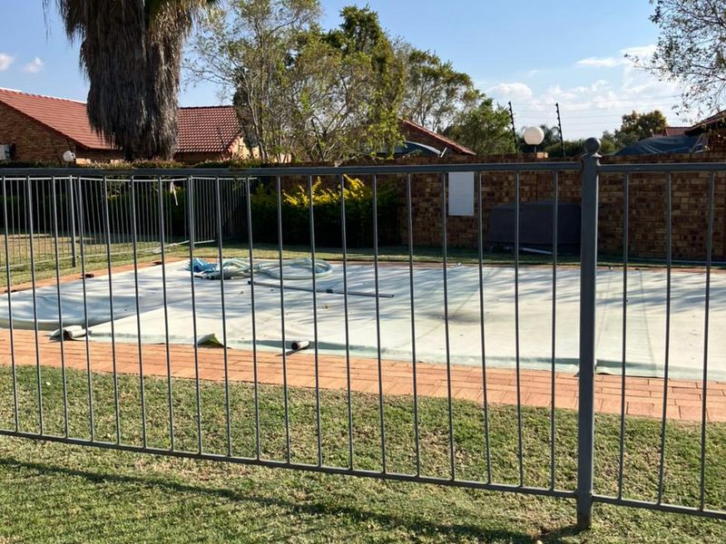 Outdoor swimming pool area featuring a protective pool cover, surrounded by brick paving and secure metal perimeter fencing within a landscaped garden setting.