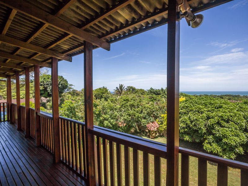 A spacious covered timber balcony featuring a wooden balustrade and exposed rafters, offering elevated views over the lush garden and distant ocean.