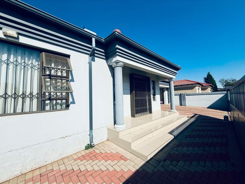 Front entrance area featuring tiled steps, decorative columns, and secure window bars, set against a paved driveway and boundary wall.