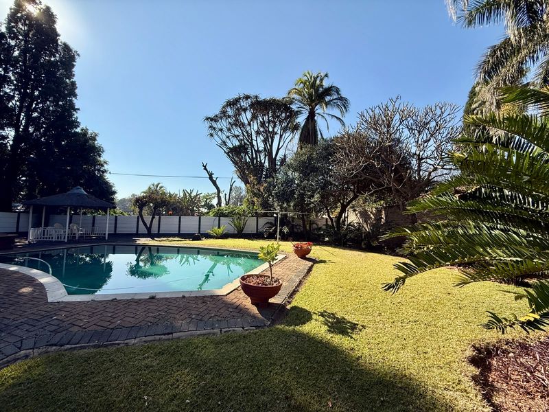 A well-maintained outdoor pool area featuring a swimming pool, brick paved surround, established garden, and a thatched gazebo.