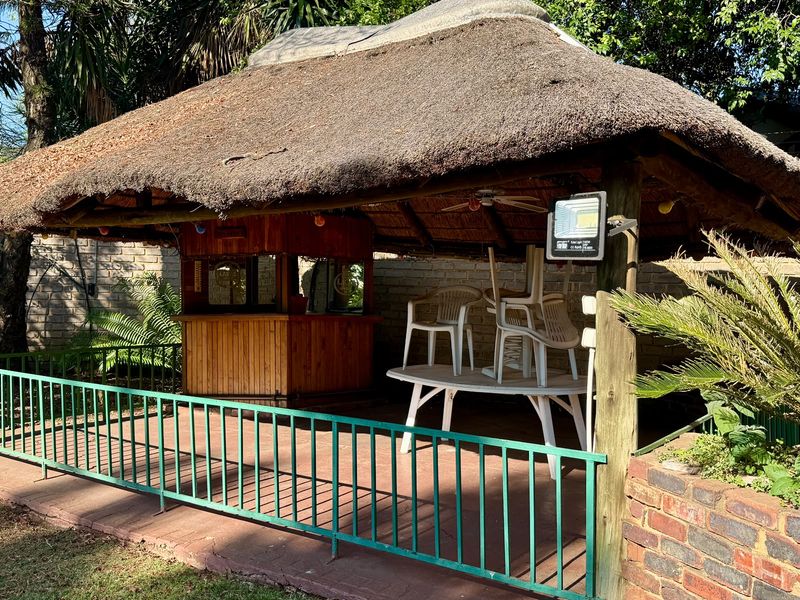 Outdoor thatched lapa area featuring a built-in timber bar counter, paved flooring, and perimeter metal railing, ideal for garden entertainment.