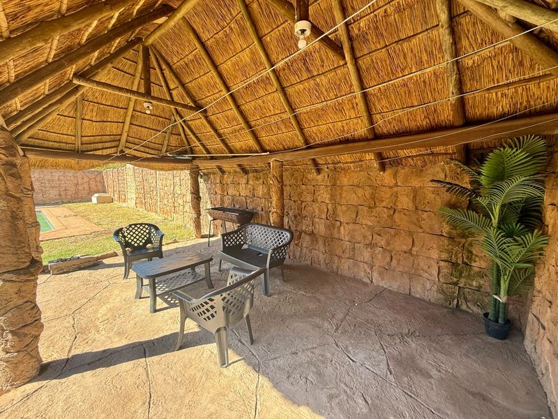 Covered outdoor patio area featuring a traditional thatched roof, exposed timber beams, and rustic stone wall cladding, overlooking the garden and pool area.