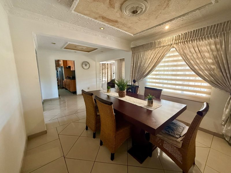 Dining room featuring tiled floors, decorative coffered ceilings with recessed lighting, and large windows with blinds, leading through to the kitchen area.