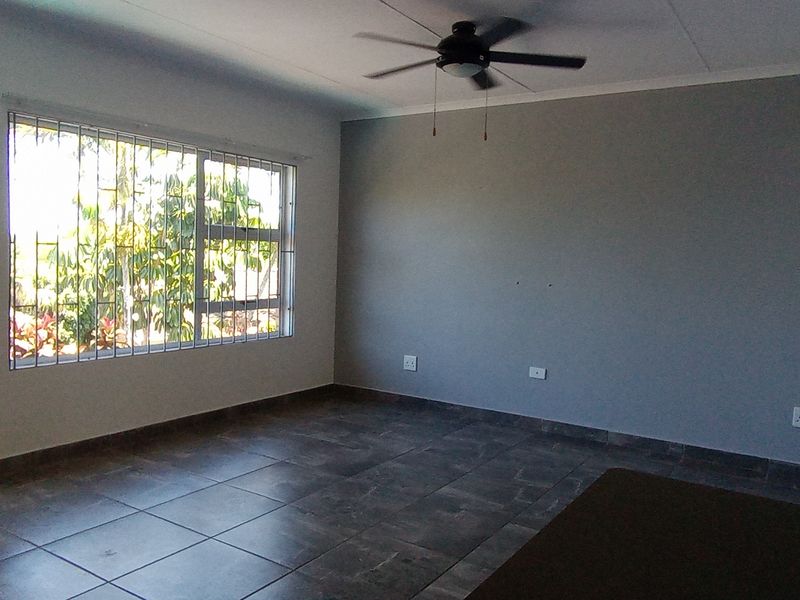 An empty living room featuring dark tiled flooring, a ceiling fan, and large windows with security bars, leading out to an exterior space.