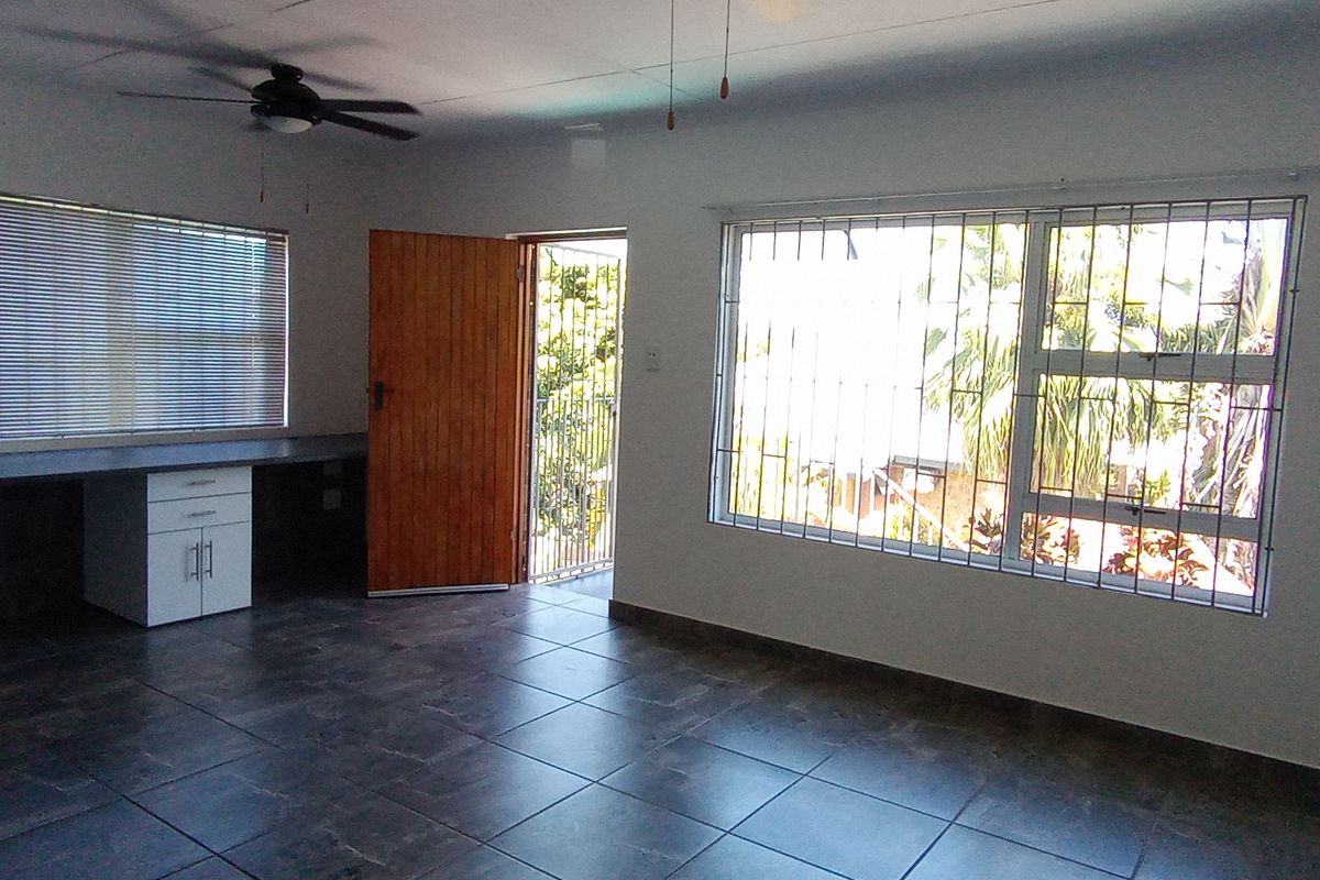An open-plan living area featuring dark tiled flooring, a built-in desk, ceiling fan, and large windows with security bars.