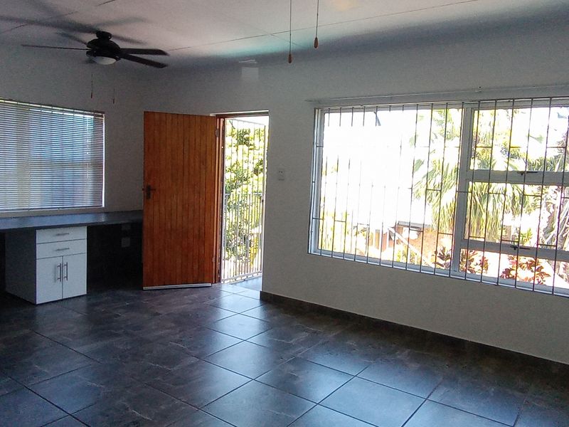 An open-plan living area featuring dark tiled flooring, a built-in desk, ceiling fan, and large windows with security bars.