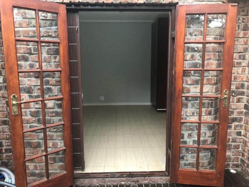 An empty bedroom viewed through open timber french doors, featuring tiled flooring and a built-in cupboard, set within an exposed brick exterior wall.