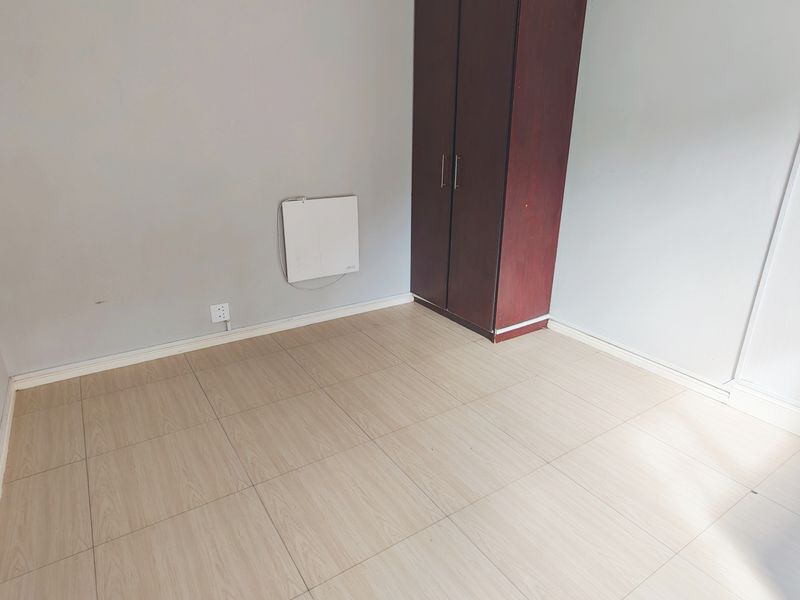 A bright, empty bedroom featuring light wood-look tiled flooring, a built-in wardrobe, and a wall-mounted heater.