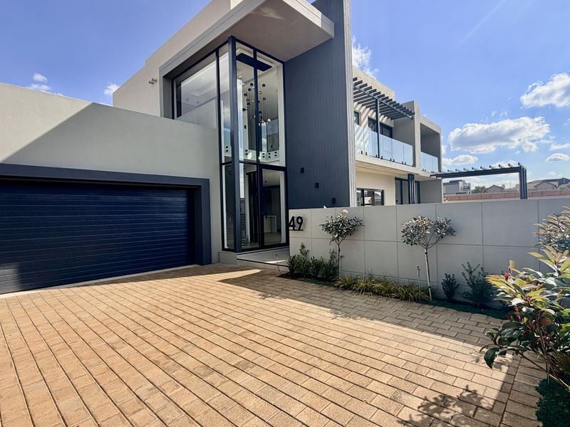 Modern double-story residential facade featuring a paved driveway, double garage, and striking full-height glass entrance with contemporary architectural detailing.