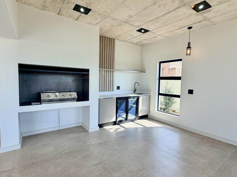 Modern kitchen area featuring an exposed concrete ceiling, built-in braai, sleek cabinetry, and high-quality tiled flooring in a contemporary open-plan space.