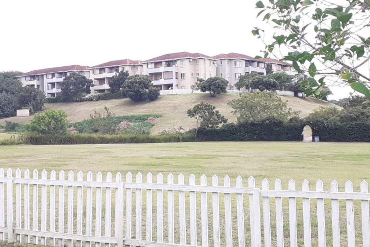 A view of a multi-story apartment complex situated on a hill, overlooking a large grassy field enclosed by a white picket fence.