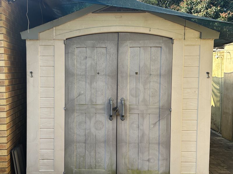 A detached prefabricated storage shed located on a paved area, featuring double doors and an overhead shade cloth structure.