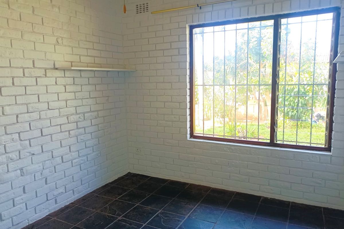 A bright, empty bedroom featuring painted white brick walls, dark slate tile flooring, and a window fitted with security bars.