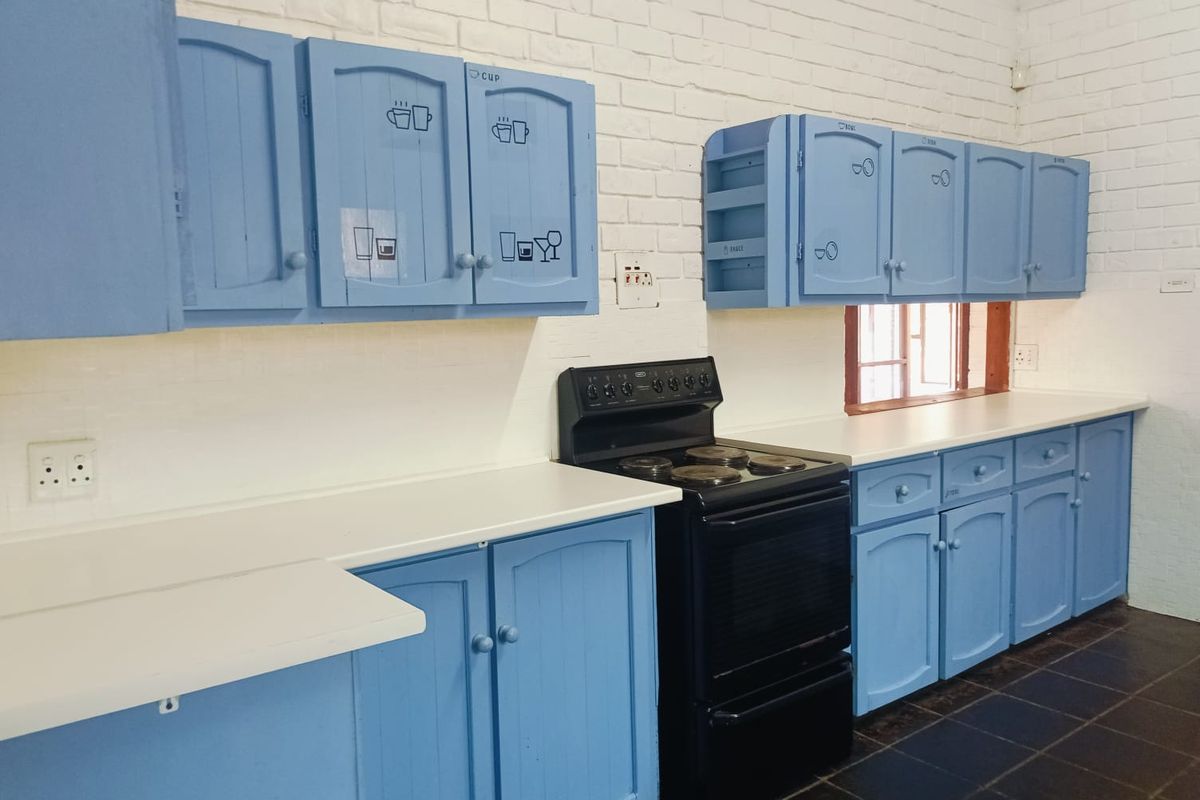 A functional kitchen featuring blue built-in cabinetry, a black electric stove, white laminate countertops, and exposed brick walls with dark tiled flooring.
