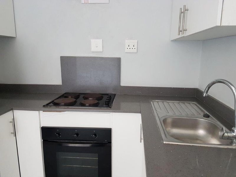 A compact kitchen featuring white cabinetry, stone countertops, a built-in oven, electric hob, and a stainless steel sink.