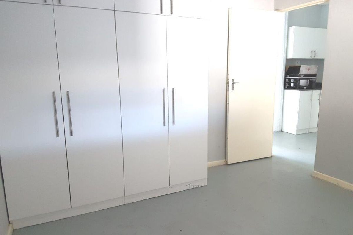A clean, empty bedroom featuring white built-in cupboards and neutral flooring, with a view through to an adjacent kitchen area.
