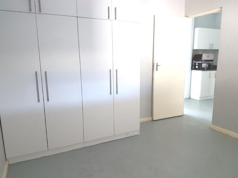 A clean, empty bedroom featuring white built-in cupboards and neutral flooring, with a view through to an adjacent kitchen area.
