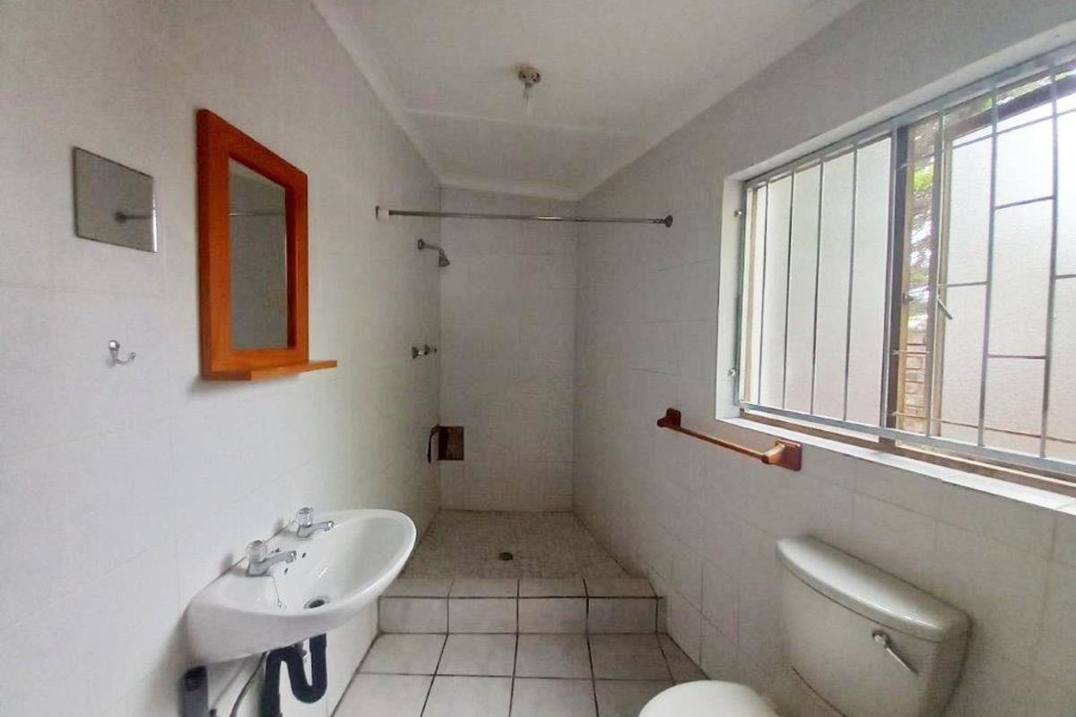 A functional bathroom featuring a walk-in shower, pedestal basin, and toilet, with tiled finishes and security bars on the window.