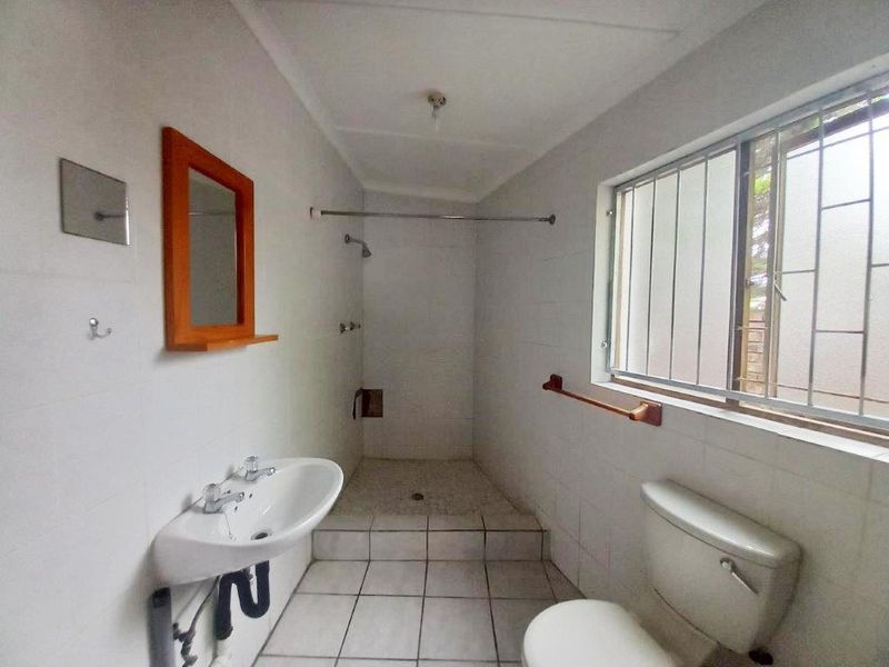 A functional bathroom featuring a walk-in shower, pedestal basin, and toilet, with tiled finishes and security bars on the window.