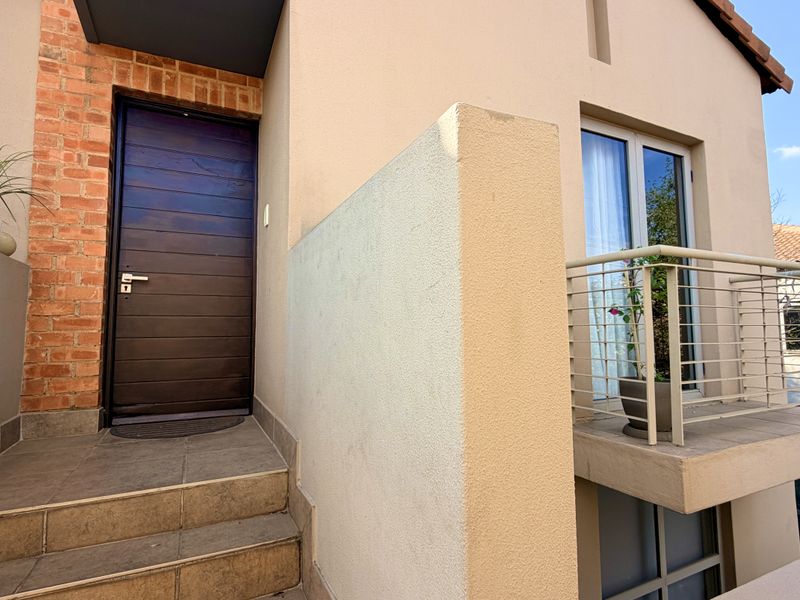Entrance area featuring a modern dark wood door, tiled steps, exposed brick accents, and an adjacent balcony with a metal railing.