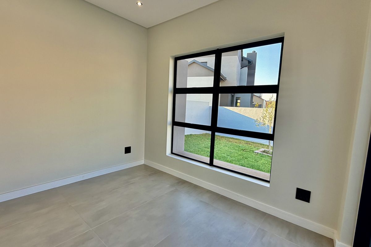 A bright, empty bedroom featuring modern large-format tiled flooring, neutral walls, and a large black-framed window overlooking the garden.