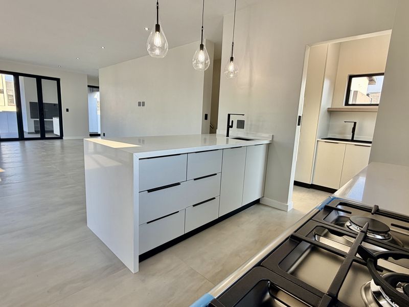 Modern open-plan kitchen featuring a central island, stone countertops, gas cooktop, and an adjacent scullery, flowing into a spacious living area with glass doors.