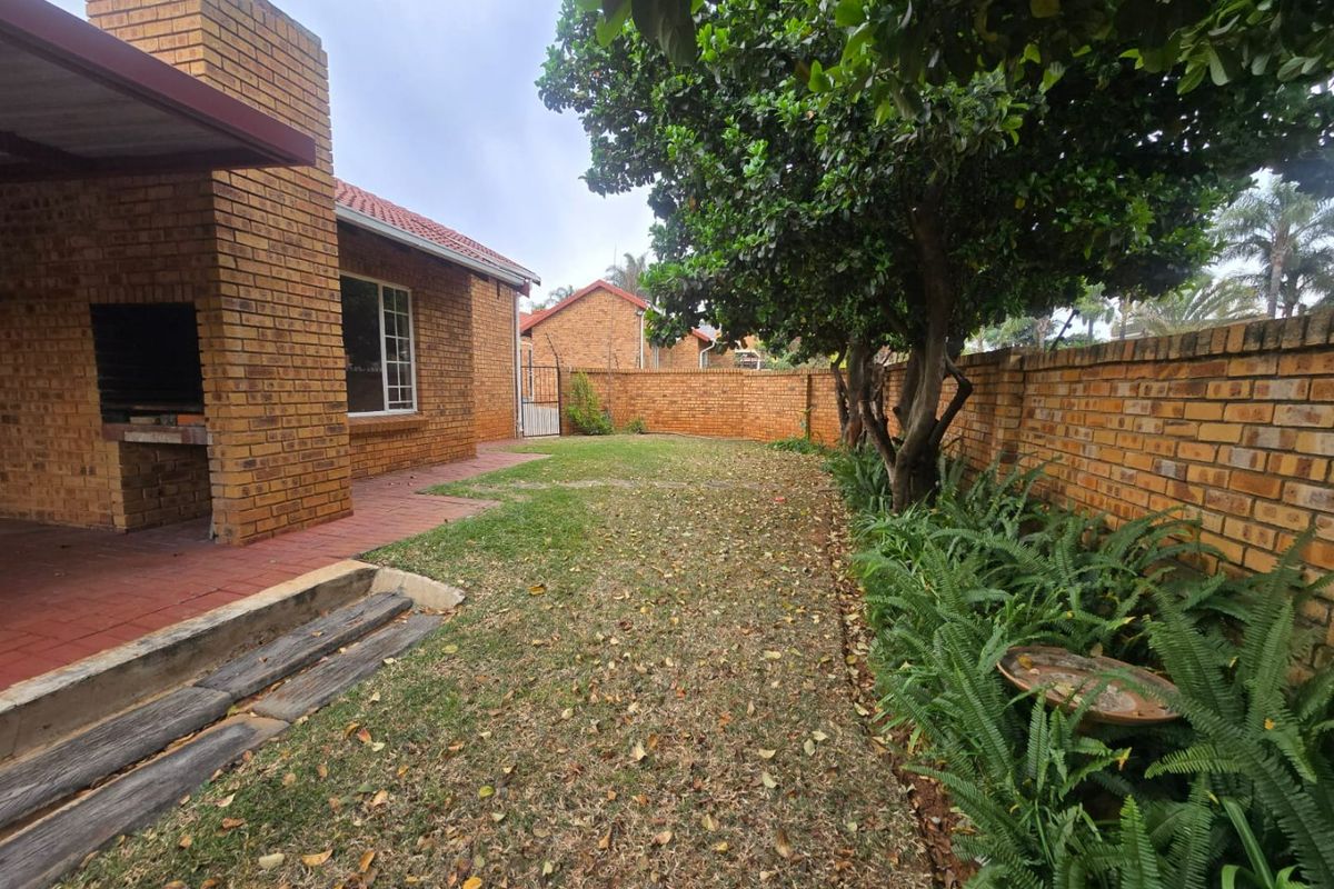 A private rear garden featuring a paved patio area with a built-in brick braai, established trees, and a secure brick boundary wall.