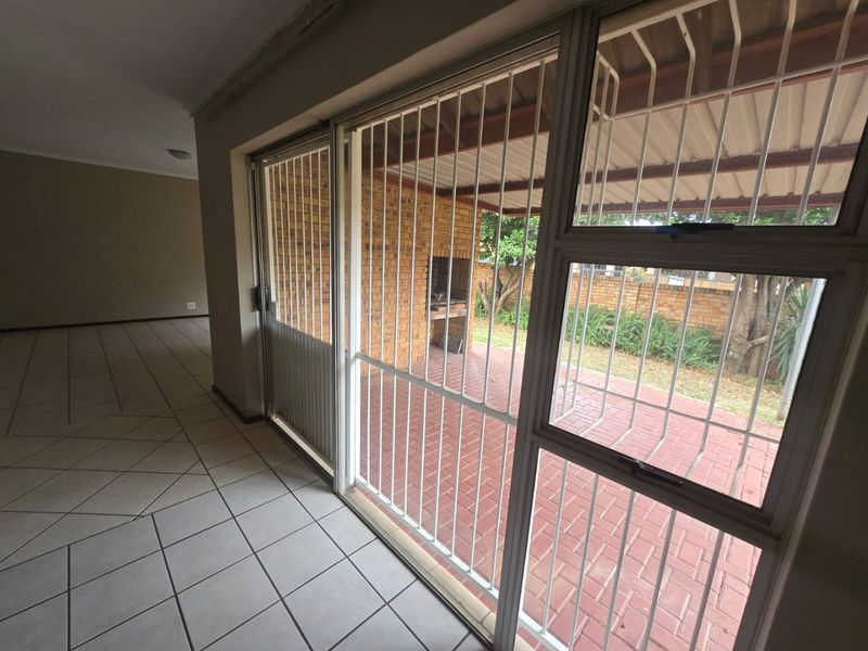 Spacious living area with tiled floors and large sliding doors opening onto a covered patio featuring a built-in braai and brick paving.