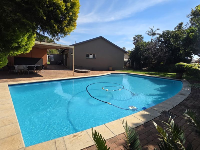 A well-maintained swimming pool area featuring a paved deck, adjacent covered patio, and established garden surroundings.