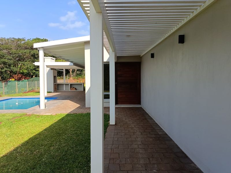A covered patio walkway featuring a pergola, leading to a timber entrance door, with views of the swimming pool and outdoor fireplace area.