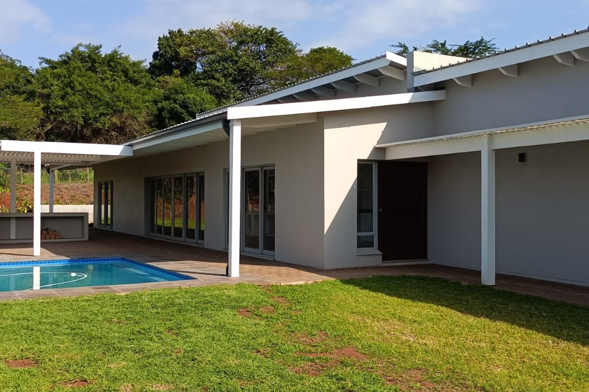 Rear exterior view featuring a modern home with a covered patio, built-in braai area, and a swimming pool set within a landscaped garden.
