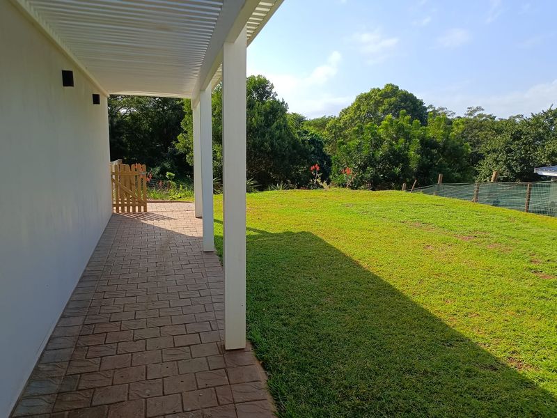 A covered paved patio area overlooking a spacious green lawn and garden, featuring structural support columns and a wooden gate.
