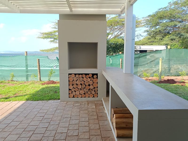 Covered outdoor patio featuring a built-in braai, concrete prep counter, and paved flooring, overlooking the garden area.