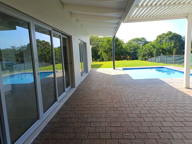 A spacious covered patio featuring paved flooring and sliding glass doors, overlooking a private swimming pool and landscaped garden area.