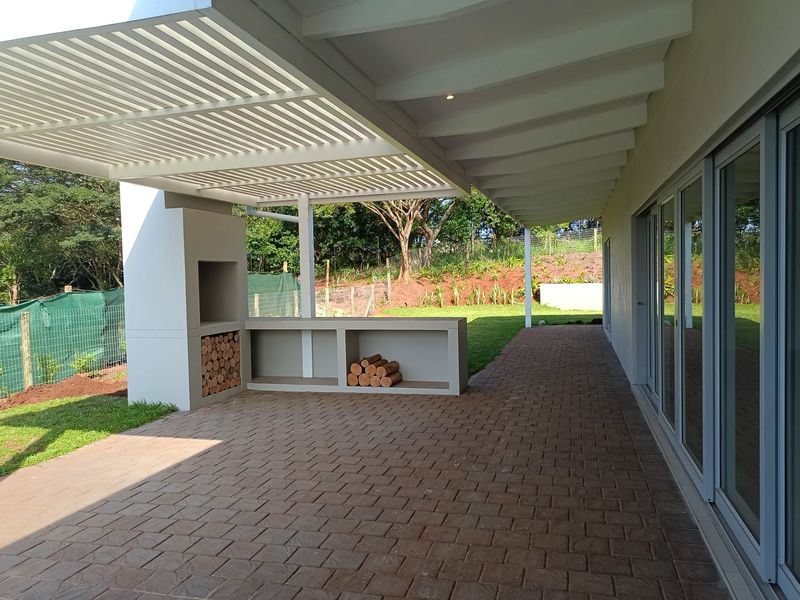 A spacious covered patio featuring a built-in braai, pergola roof, and paved flooring, providing seamless access to the garden through large sliding glass doors.