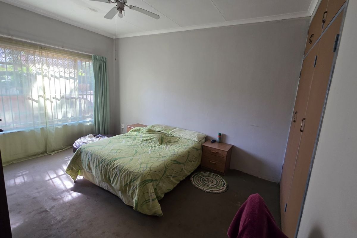 Spacious bedroom featuring built-in cupboards, a ceiling fan, and large windows with security bars, offering a functional layout in need of cosmetic updates.