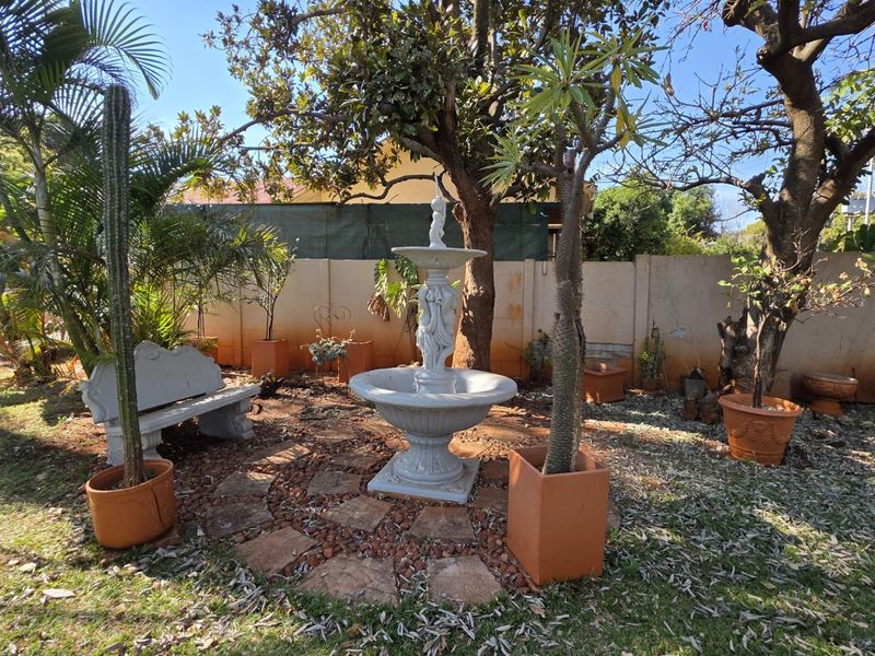 A garden area featuring a central stone fountain, stone paving, and a concrete boundary wall surrounded by mature trees and potted plants.