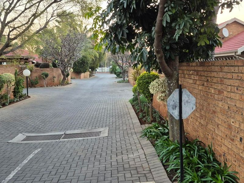 A view of the paved complex driveway lined with brick boundary walls, mature trees, and landscaping, leading towards the residential units.