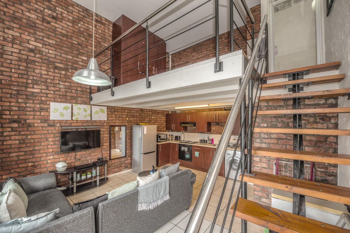 Open-plan living area featuring exposed brick walls, a mezzanine level with metal railing, and a functional kitchen with built-in cabinetry and appliances.