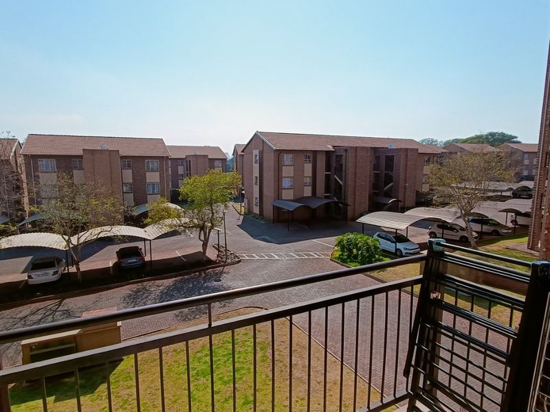 A balcony view overlooking the apartment complex grounds, featuring paved driveways, covered parking bays, and landscaped garden areas.