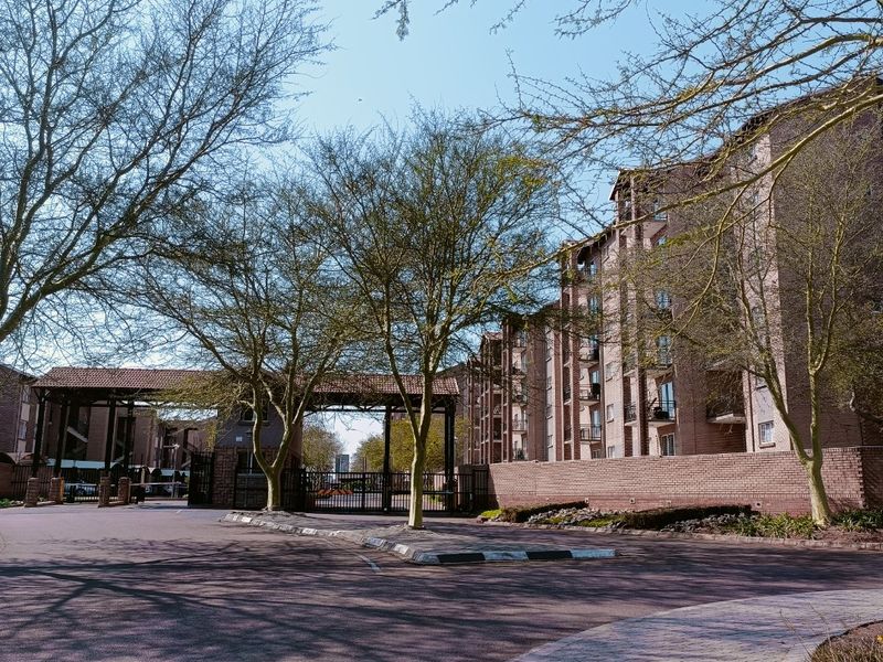 View of the apartment complex entrance featuring a brick facade, security gate, and paved driveway surrounded by mature trees.