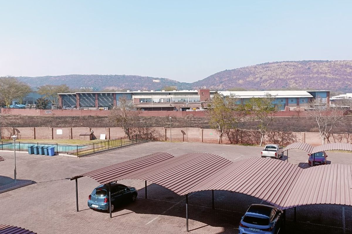 An elevated view of the apartment complex exterior, featuring paved parking areas with covered carports, perimeter walling, and a communal swimming pool.