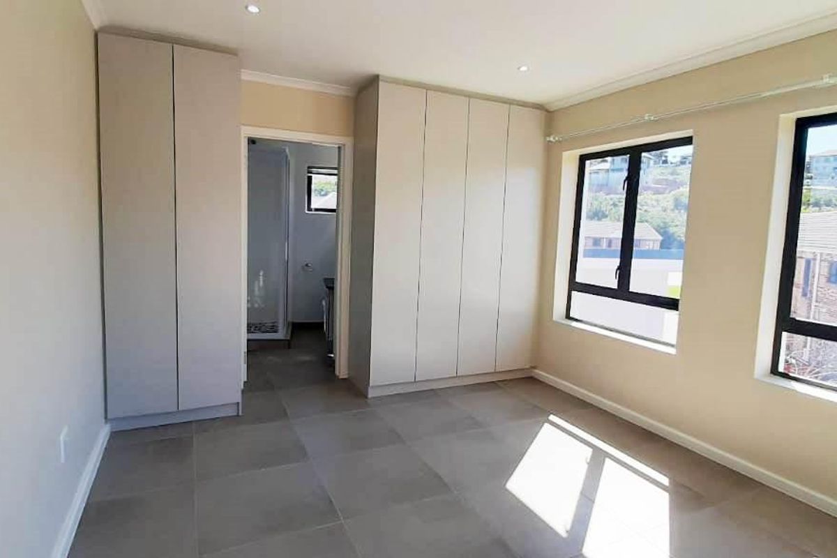 A bright, empty bedroom featuring neutral tiled flooring, built-in cupboards, and large aluminium-framed windows with views of the surrounding neighbourhood.