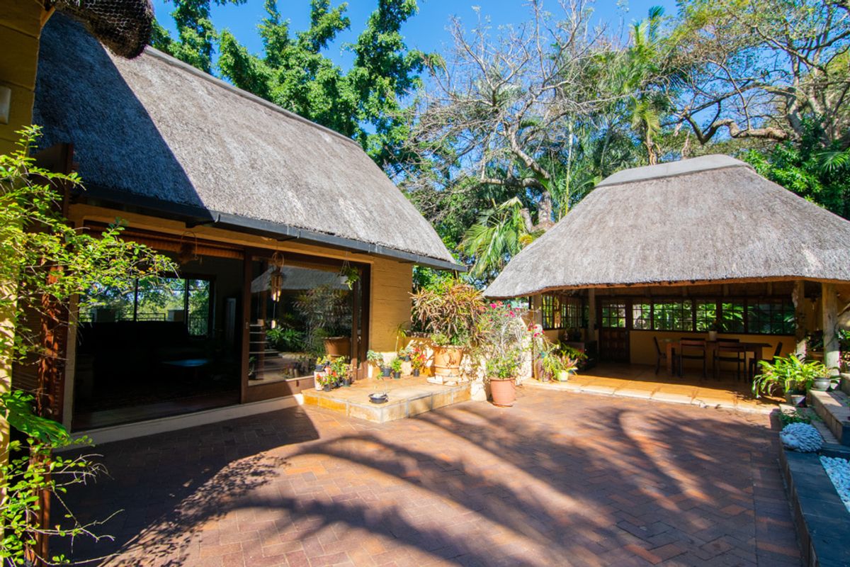 A spacious rear courtyard featuring brick paving, a covered patio area, and traditional thatch roofing, surrounded by mature trees and lush garden greenery.