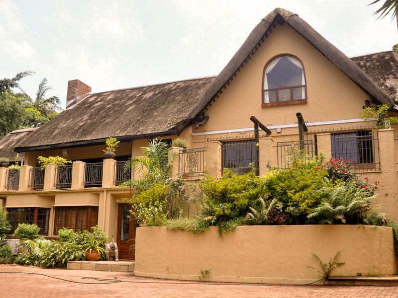 A charming multi-level home featuring a traditional thatched roof, a spacious balcony with wrought iron railings, and a paved driveway entrance.