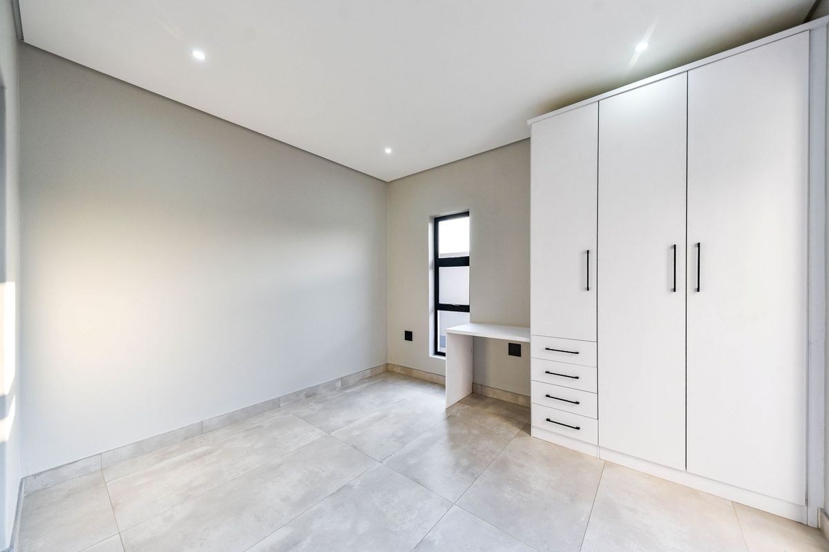 A modern, empty bedroom featuring neutral tiled flooring, recessed downlights, and a clean white built-in cupboard unit with an integrated desk.