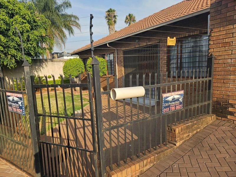 Front exterior view of a brick house featuring a paved driveway, secure metal perimeter fencing, and electric security measures.