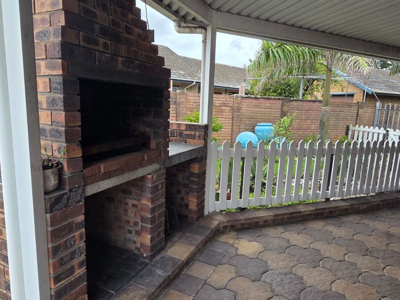 A covered outdoor patio featuring a built-in brick braai, paved flooring, and a picket fence overlooking the garden area.