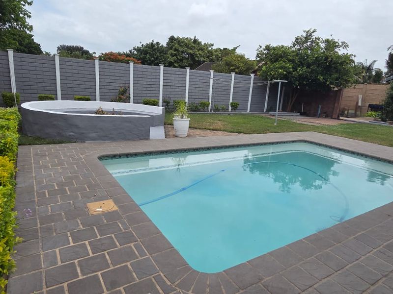 A well-maintained backyard featuring a swimming pool with paved surround, a raised circular fire pit, and a secure perimeter wall.